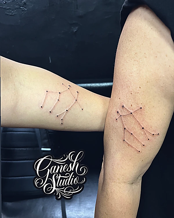 Dos brazos con la misma constelación tatuada a línea y punto, con el logo en script de Ganesh Studio en la esquina