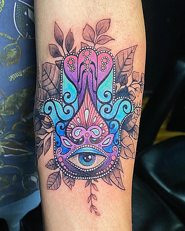 Hamsa con ojo a color en magenta, violeta y turquesa, rodeada de hojas en negro y gris, en el antebrazo