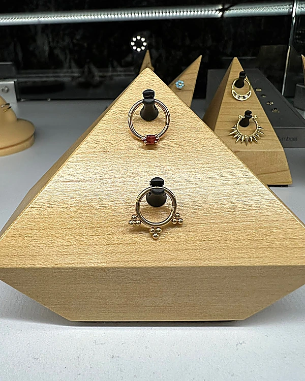 Pirámide de madera clara en la vitrina con dos aros de piercing dorados, uno con piedra roja y otro con bolitas, y más pirámides con joyería al fondo