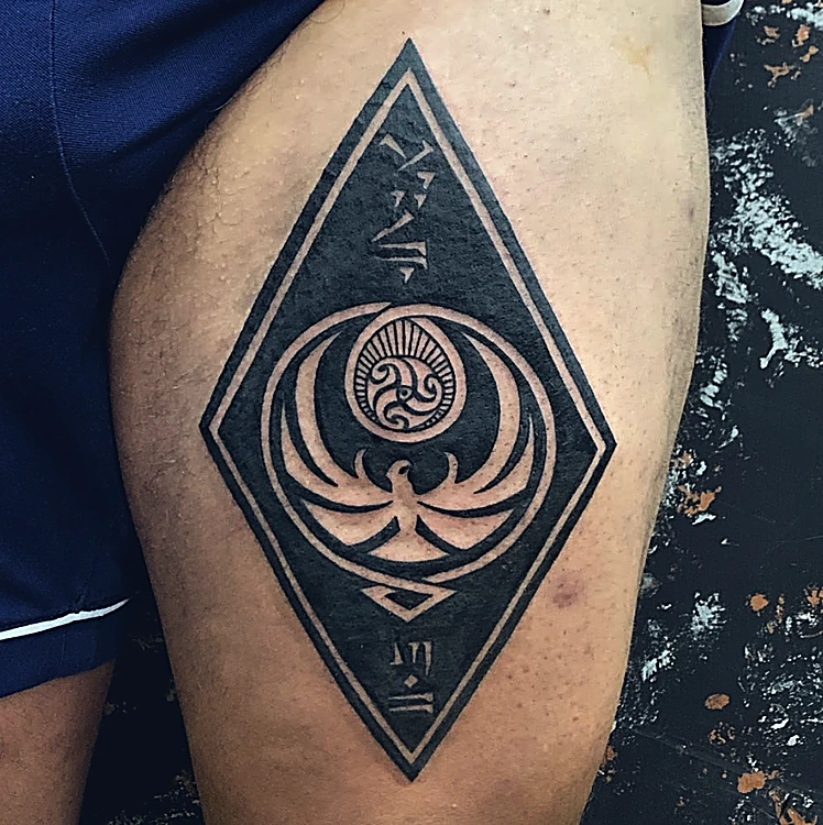Rombo de relleno negro con un ave de alas abiertas y glifos, tatuado en el muslo
